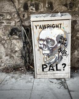 Street art on a utility cabinet. Skull says, 'Y'awright, pal?'. Someone else has replied, 'Aye, mate.'